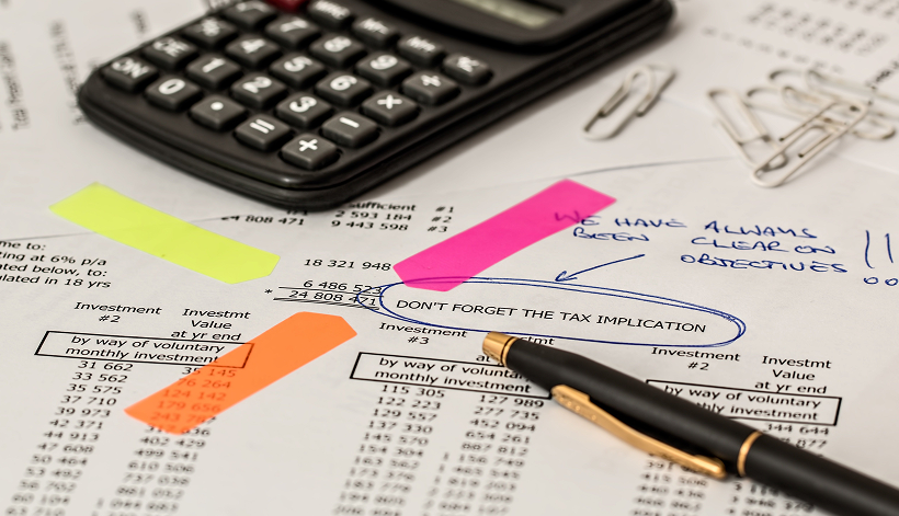 A calculator, pen, paper clips, and sheets of financial data with highlighted sections and handwritten notes, including “DON’T FORGET THE TAX IMPLICATION” and “WE HAVE ALWAYS BEEN CLEAR ON OBJECTIVES.”.
